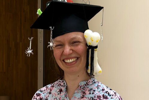 Eine lächelnde Frau trägt einen Doktorhut, der mit bunten Tierfiguren und einem Knochenspielzeug dekoriert ist. Sie trägt ein buntes Hemd und steht in einem hellen Raum. Der Hut hebt sich durch die ungewöhnliche Gestaltung hervor.