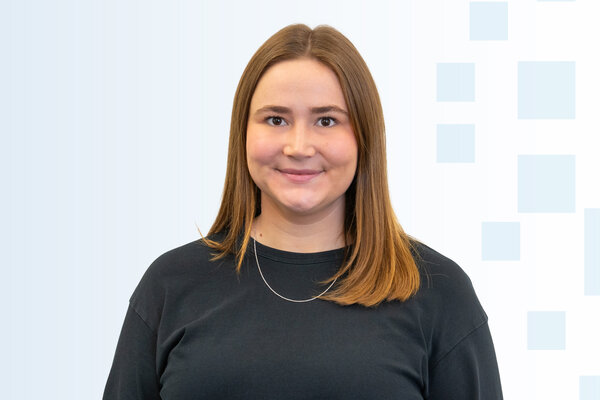 Ein lächelnde Frau mit langen, braunen Haaren trägt ein schwarzes Sweatshirt. Sie steht vor einem hellen, leicht strukturierten Hintergrund. Ihre Pose vermittelt Freundlichkeit und Offenheit.