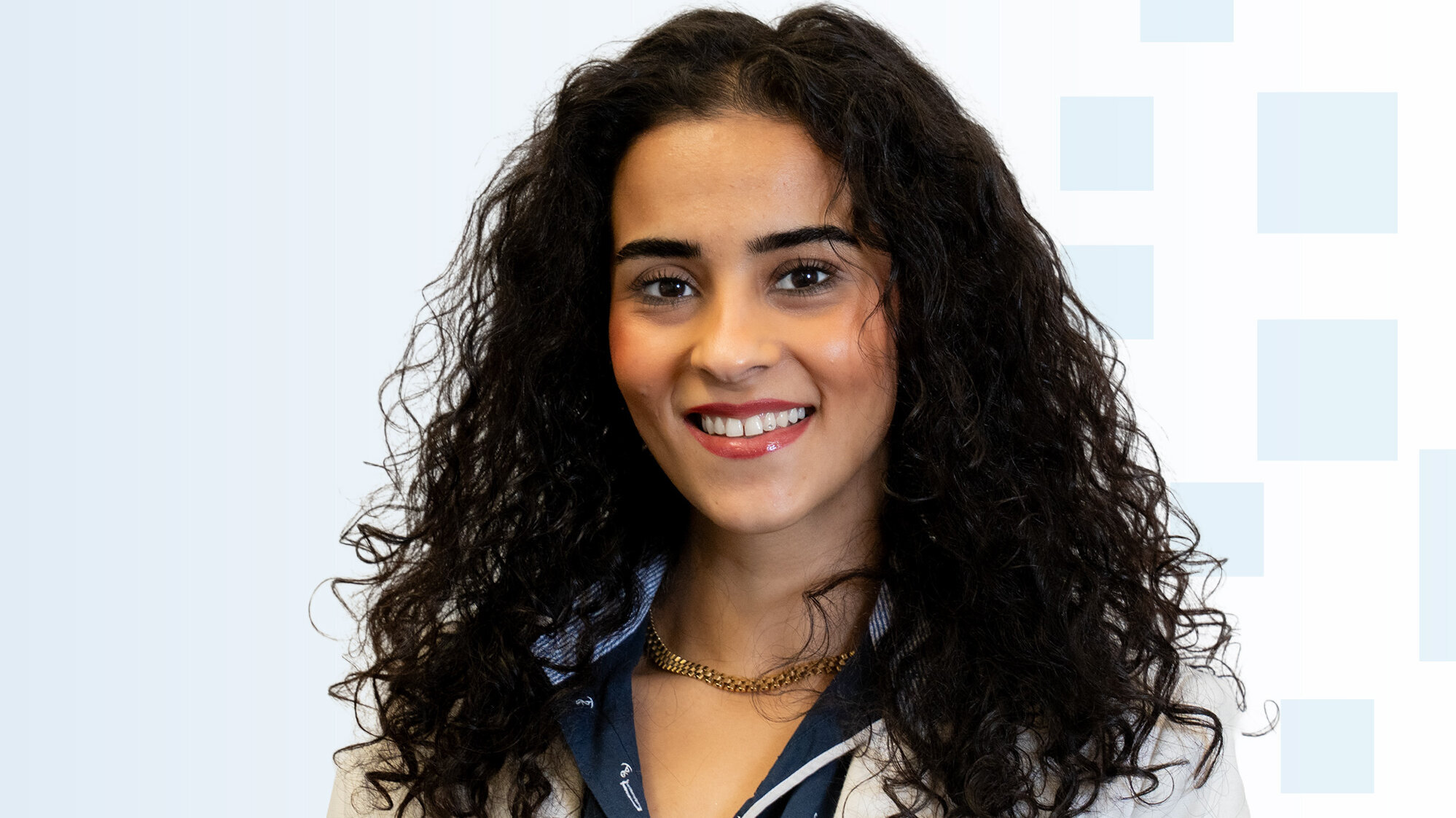 Ein Porträt einer lächelnden jungen Frau mit lockigem, dunklem Haar. Sie trägt ein helles Jackett und eine blaue Bluse. Der Hintergrund ist hell und zeigt sanfte geometrische Formen.