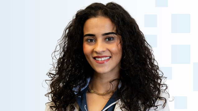 Ein Porträt einer lächelnden jungen Frau mit lockigem, dunklem Haar. Sie trägt ein helles Jackett und eine blaue Bluse. Der Hintergrund ist hell und zeigt sanfte geometrische Formen.