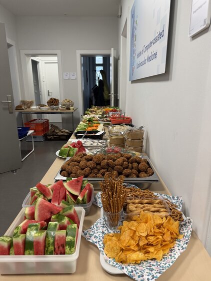 Ein langer Tisch ist mit verschiedenen Snacks gedeckt. Darauf liegen Wassermelonenstücke, Snacks wie Chips und Brezeln sowie süße und salzige Häppchen. Im Hintergrund sind weitere Speisen und eine hell gestrichene Wand zu sehen.