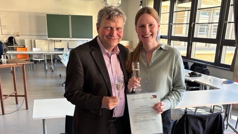Ein Mann und eine Frau stehen in einem Raum, beide halten ein Glas. Die Frau zeigt ein Diplom oder Zertifikat in der Hand. Sie lächeln, während sie nebeneinander posieren. Im Hintergrund sind Tische und Stühle sichtbar.