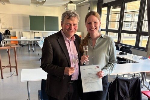 Ein Mann und eine Frau stehen in einem Raum, beide halten ein Glas. Die Frau zeigt ein Diplom oder Zertifikat in der Hand. Sie lächeln, während sie nebeneinander posieren. Im Hintergrund sind Tische und Stühle sichtbar.