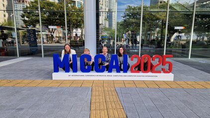 Vier Frauen posieren vor einem großen Schriftzug „MICCAI 2025“ in den Farben blau und rot. Der Schriftzug steht für die 26. internationale Konferenz zu medizinischer Bildverarbeitung und computergestützter Intervention. Im Hintergrund sind moderne Gebäude und Bäume sichtbar.