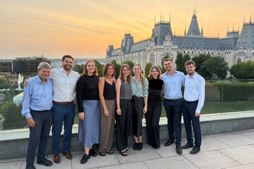 Eine Gruppe von neun Personen steht vor einem schönen historischen Gebäude während des Sonnenuntergangs. Sie lächeln und tragen unterschiedliche, elegante Kleidung. Der Hintergrund zeigt eine grüne Landschaft und die Silhouette des Gebäudes wird durch das warme Licht des Himmels hervorgehoben.