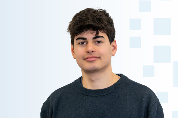 Ein junger Mann mit dunklen lockigen Haaren trägt einen schwarzen Pullover. Er blickt direkt in die Kamera vor einem hellen, dezenten Hintergrund mit geometrischen Formen.