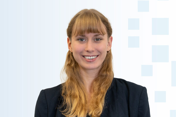 Eine junge Frau mit langen, blonden Haaren und einladendem Lächeln trägt einen dunklen Blazer. Der Hintergrund ist hell und enthält geometrische Formen in unterschiedlichen Blau-Tönen. Sie wirkt freundlich und professionell.