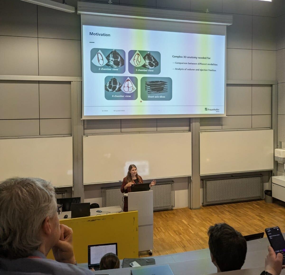 Eine Frau steht vor einem Präsentationsbildschirm in einem Hörsaal und erklärt Inhalte. Der Bildschirm zeigt verschiedene Diagramme und Texte mit dem Titel "Motivationen". Im Vordergrund sind teilweise der Kopf und die Schultern von Zuhörern zu sehen.