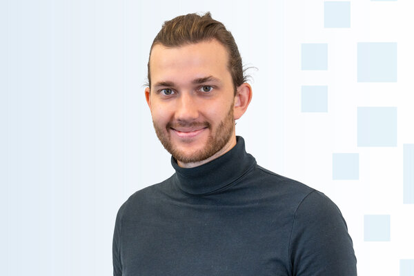 Ein junger Mann mit einem lächelnden Gesicht trägt ein schwarzes Rollkragenoberteil. Der Hintergrund ist hell und zeigt leichte geometrische Muster.