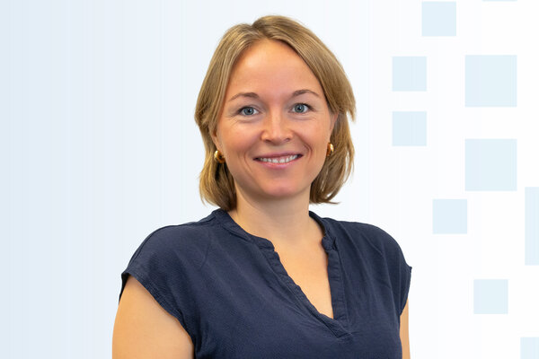 Porträt einer lächelnden Frau mit blonden Haaren, die ein schwarzes Oberteil trägt. Der Hintergrund ist hell und hat geometrische Formen. Sie wirkt freundlich und zugänglich.