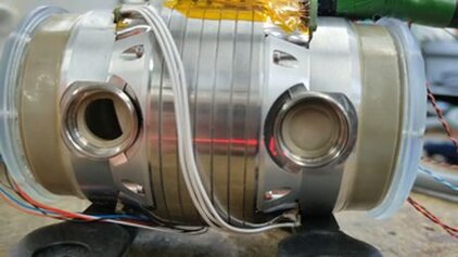 Das Bild zeigt einen metallischen Zylinder mit zwei Öffnungen an den Seiten und verschiedenen Kabelverbindungen, die daran befestigt sind. Die Oberfläche ist glänzend und silbern, mit sichtbaren Schichten und Drähten, die an die Struktur angeschlossen sind.