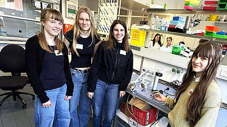 Vier junge Frauen stehen in einem Labor, lächeln und posieren für das Foto. Sie tragen lässige Kleidung und Namensschilder. Im Hintergrund sind Labormaterialien und Geräte sichtbar. Der Raum wirkt modern und gut ausgestattet.