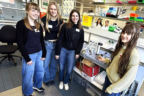 Vier junge Frauen stehen in einem Labor, lächeln und posieren für das Foto. Sie tragen lässige Kleidung und Namensschilder. Im Hintergrund sind Labormaterialien und Geräte sichtbar. Der Raum wirkt modern und gut ausgestattet.