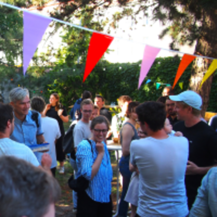 Eine fröhliche Gartenparty mit verschiedenen Menschen, die miteinander plaudern und lachen. Bunte Wimpel hängen über der Veranstaltung. Die Atmosphäre ist entspannt und gesellig, mit Gästen, die sich in Gruppen unterhalten und den Anlass genießen.