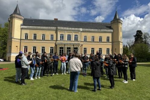 Eine Gruppe von etwa 20 Personen steht im Freien auf einer Wiese vor einem großen, gelben Schlossgebäude. Die Menschen wirken beschäftigt, einige halten Notizen oder Liederblätter in der Hand. Hinter ihnen sind Bäume und ein bewölkter Himmel sichtbar.
