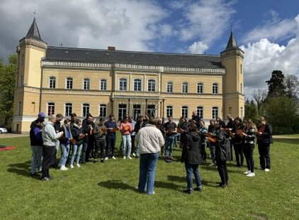 Eine Gruppe von etwa 20 Personen steht im Freien auf einer Wiese vor einem großen, gelben Schlossgebäude. Die Menschen wirken beschäftigt, einige halten Notizen oder Liederblätter in der Hand. Hinter ihnen sind Bäume und ein bewölkter Himmel sichtbar.