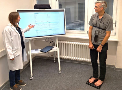 Eine Person in einem Laborkittel zeigt auf einen Bildschirm, auf dem Daten angezeigt werden. Neben ihr steht ein Mann barfuß auf einer Waage. Der Bildschirm zeigt verschiedene Messwerte und Grafiken.