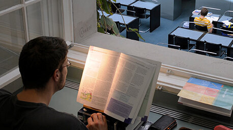 Ein Mann sitzt an einem Tisch und studiert ein Buch, das im Licht einer Fensterscheibe aufgeblättert ist. Im Hintergrund sind Tische und Stühle zu sehen, wo weitere Personen in einer Bibliothek oder einem Lernraum sitzen.