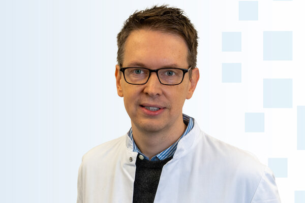 Ein Arzt trägt einen weißen Kittel und eine Brille. Er steht vor einem hellen, grafischen Hintergrund. Der Fokus liegt auf seinem freundlichen Gesichtsausdruck und seiner professionellen Ausstrahlung.