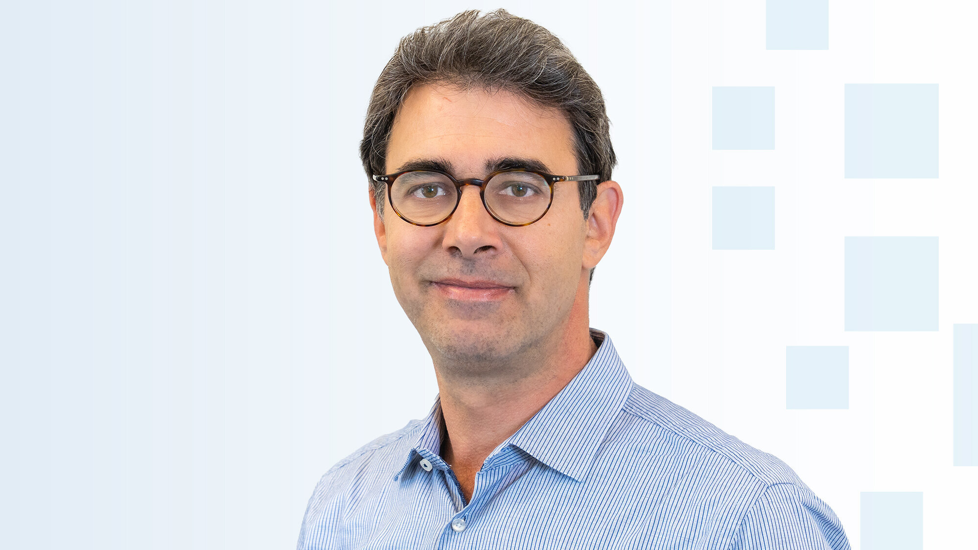 Ein Mann mit bräunlichen Haaren trägt eine Brille und einen blauen Anzug. Er hat ein freundliches Lächeln und steht vor einem hellen, geometrischen Hintergrund.