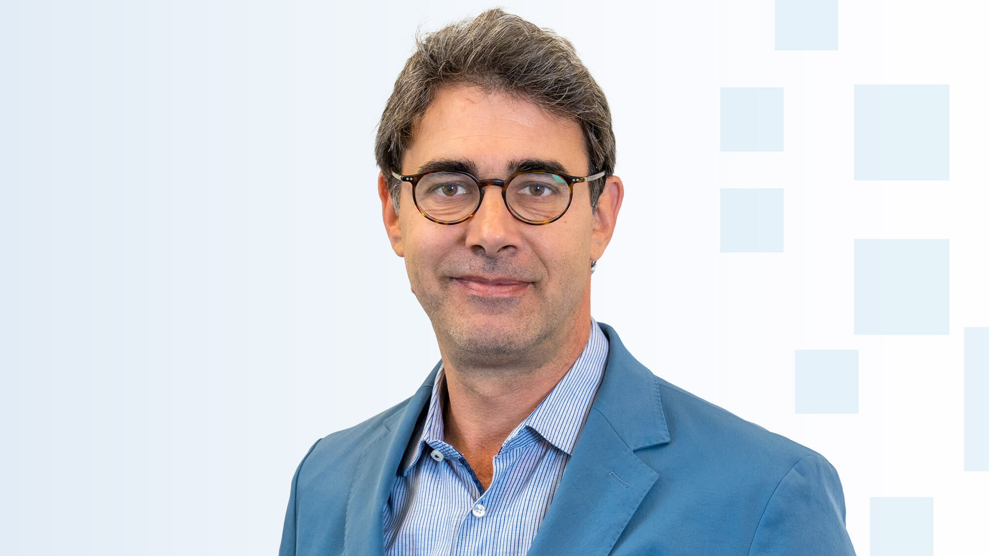 Ein Mann mit bräunlichen Haaren trägt eine Brille und einen blauen Anzug. Er hat ein freundliches Lächeln und steht vor einem hellen, geometrischen Hintergrund.