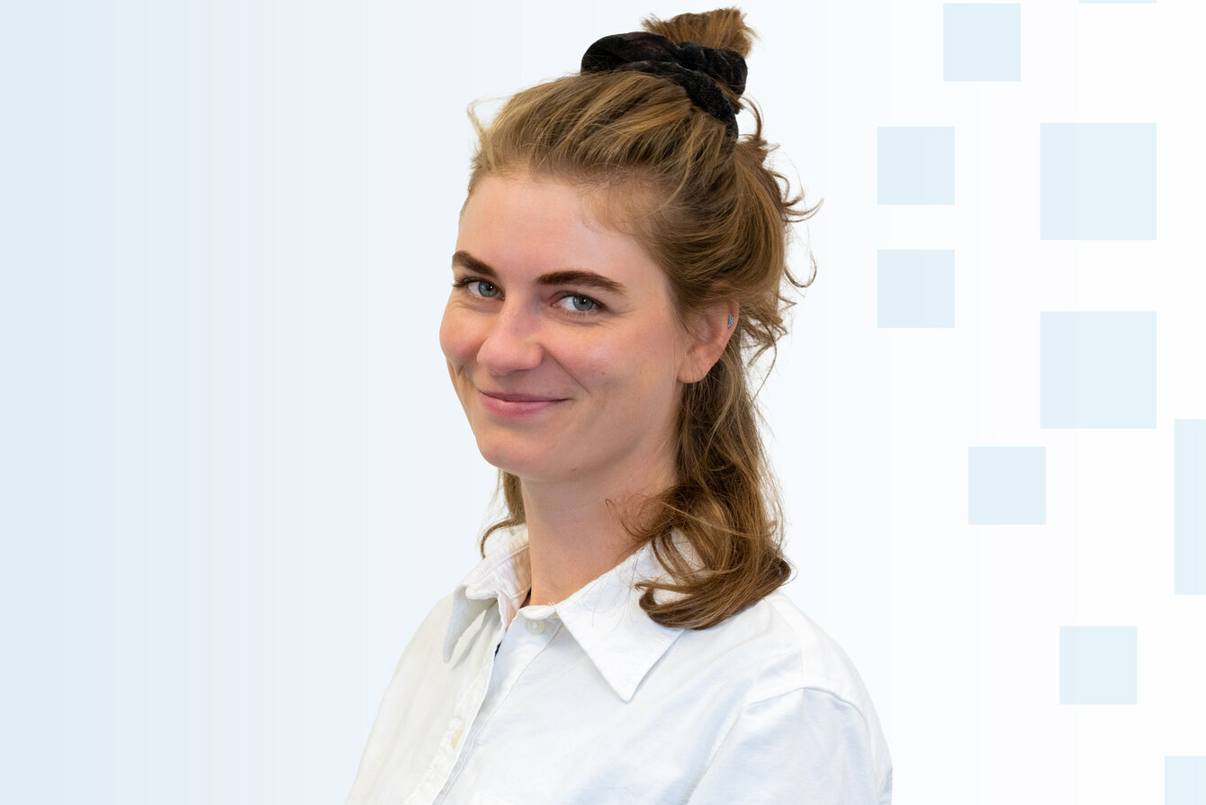 Eine junge Frau mit Locken, die zu einem halben Pferdeschwanz gebunden sind, lächelt freundlich. Sie trägt ein weißes Hemd. Der Hintergrund ist hell und enthält geometrische Formen in sanften Farben.