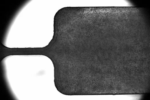 Das Bild zeigt eine stark vergrößerte und kontrastreiche Darstellung eines metallischen Objekts mit einer rechteckigen Form und einem schmalen Ansatz. Die Oberfläche ist rau und hat eine matte Textur, während der Hintergrund hell und unscharf ist.