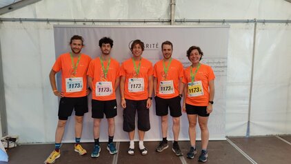 Fünf Personen in orangefarbenen T-Shirts stehen lächelnd vor einem Hintergrund mit einem Logo. Alle tragen Medaillen um den Hals und haben die Startnummer 1173. Sie scheinen nach einerSportveranstaltung posieren.