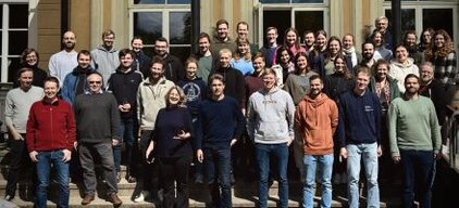 Gruppenfoto von etwa 50 Personen, die vor einer großen Fensterfront stehen. Die Teilnehmer lächeln und sind in alltäglicher Kleidung. Die Aufnahme vermittelt eine freundliche, gemeinschaftliche Atmosphäre. Es handelt sich um eine gemischte Gruppe aus Männern und Frauen unterschiedlichen Alters.