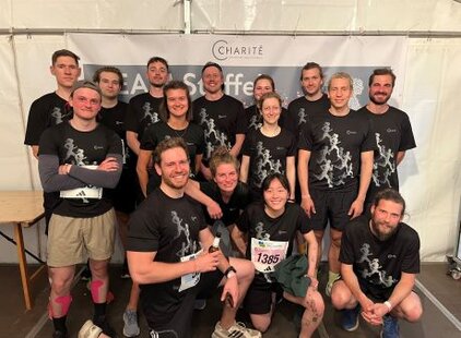 Eine Gruppe von 14 Personen steht in Sportbekleidung vor einem Banner der Charité. Die Teilnehmer lächeln und posieren gemeinschaftlich für das Foto, während einige sportliche Accessoires sichtbar sind. Die Atmosphäre wirkt freundlich und motiviert.