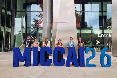 Sechs Personen posieren vor einem modernen Gebäude. Sie stehen hinter großen, blauen Buchstaben, die "MICCAI 26" bilden. Die Szene vermittelt ein Gefühl der Begeisterung und Zusammengehörigkeit, wahrscheinlich im Rahmen einer Konferenz oder Veranstaltung.