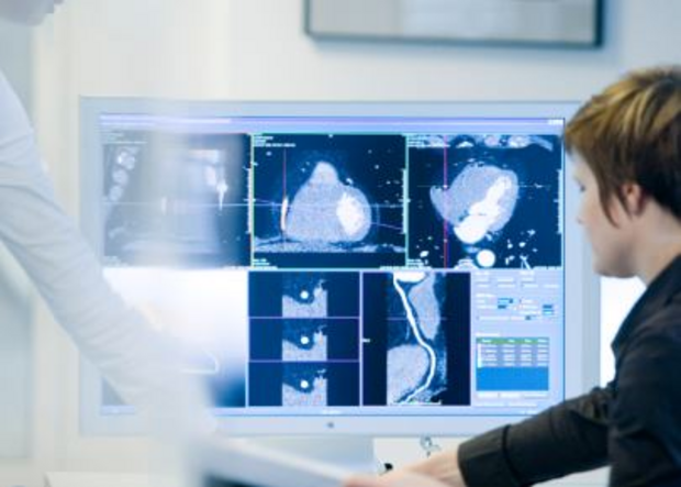 Eine Person sitzt vor einem Computerbildschirm, auf dem mehrere medizinische Bildgebungen wie CT-Scans zu sehen sind. Eine zweite Person zeigt auf den Bildschirm. Die Szene vermittelt den Austausch von Informationen im medizinischen Kontext.