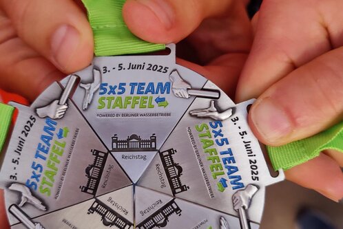 Sechs Hände halten eine Medaille für die „5x5 Teamstaffel“, die vom 3. bis 5. Juni 2025 stattfindet. Die Medaille hat eine runde Form und trägt den Schriftzug der Veranstaltung sowie ein abgebildetes Gebäude.