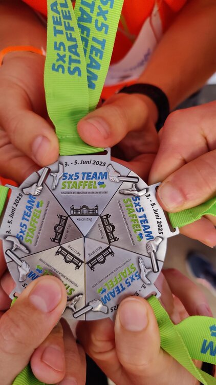 Sechs Hände halten eine Medaille für die „5x5 Teamstaffel“, die vom 3. bis 5. Juni 2025 stattfindet. Die Medaille hat eine runde Form und trägt den Schriftzug der Veranstaltung sowie ein abgebildetes Gebäude.