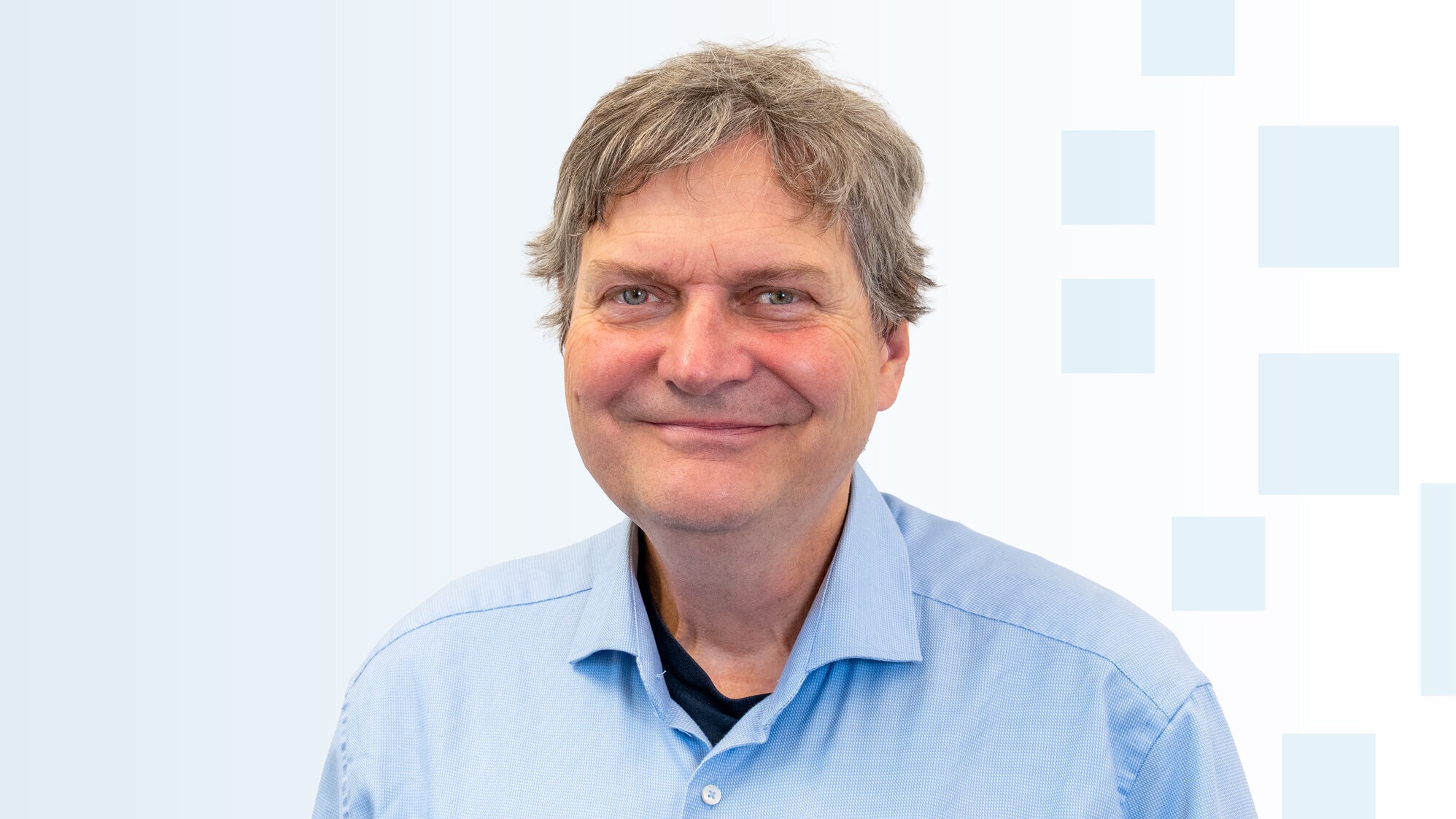 Ein lächelnder Mann mit schulterlangen, grauen Haaren trägt ein hellblaues Hemd. Er steht vor einem sanften, hellen Hintergrund mit geometrischen Formen. Der Mann strahlt Freundlichkeit aus.
