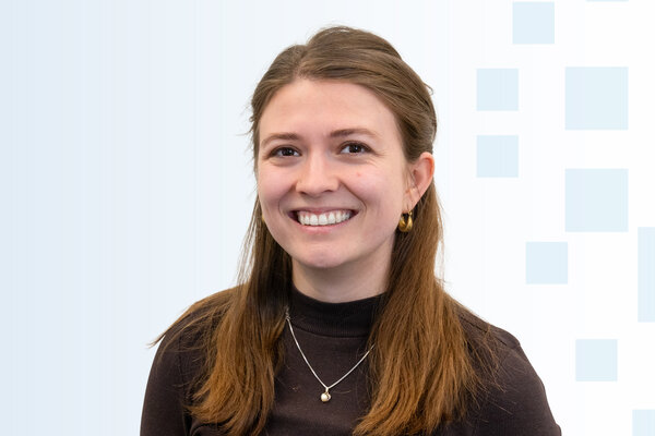 Ein junger Mann mit langen braunen Haaren lächelt. Er trägt ein dunkles Oberteil und eine dezente Halskette. Der Hintergrund ist hell und enthält geometrische Formen in einem sanften Farbton.