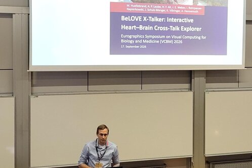 Ein Mann steht an einem Podium und präsentiert einen Vortrag. Im Hintergrund ist eine Folie mit dem Titel "BeLOVE X-Talker: Interactive Heart-Brain Cross-Talk Explorer" und Informationen zu einem Symposium über visuelle Computation für Biologie und Medizin zu sehen.