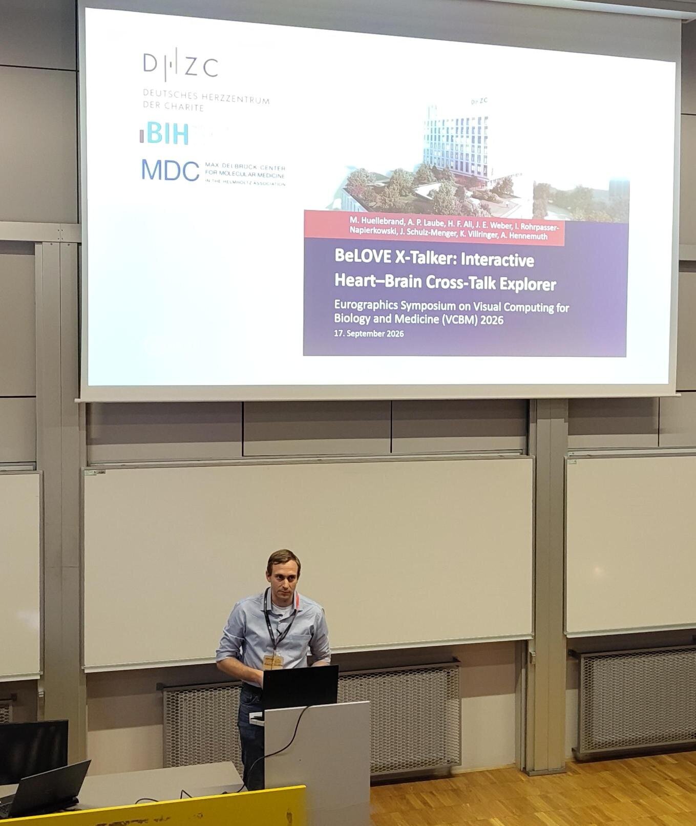 Ein Mann steht an einem Podium und präsentiert einen Vortrag. Im Hintergrund ist eine Folie mit dem Titel "BeLOVE X-Talker: Interactive Heart-Brain Cross-Talk Explorer" und Informationen zu einem Symposium über visuelle Computation für Biologie und Medizin zu sehen.