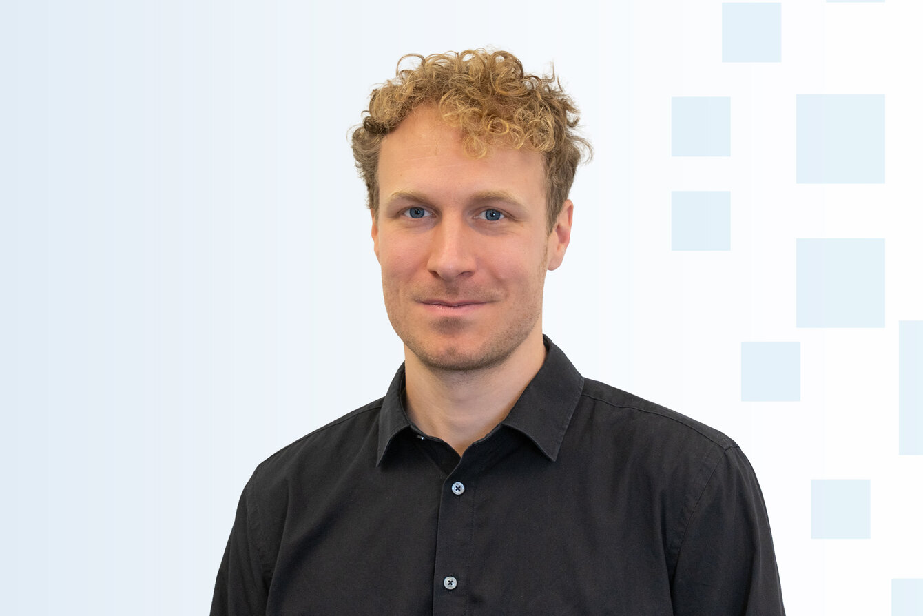 Ein Mann mit lockigem, blonden Haar trägt ein schwarzes Hemd. Er hat einen neutralen Gesichtsausdruck und steht vor einem hellen, strukturierten Hintergrund.