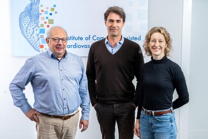 Das Bild zeigt drei Personen, die zusammenstehen. Links ein älterer Mann mit Brille und grauem Haar, in einem hellblauen Hemd. In der Mitte ein jüngerer Mann mit dunklem Haar, der ein schwarzes Pullover trägt. Rechts eine junge Frau mit lockigem Haar, die einen schwarzen Rollkragenpullover anhat. Der Hintergrund zeigt ein Banner des Instituts.