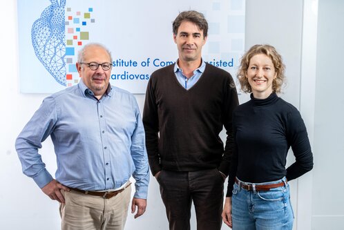 Das Bild zeigt drei Personen, die zusammenstehen. Links ein älterer Mann mit Brille und grauem Haar, in einem hellblauen Hemd. In der Mitte ein jüngerer Mann mit dunklem Haar, der ein schwarzes Pullover trägt. Rechts eine junge Frau mit lockigem Haar, die einen schwarzen Rollkragenpullover anhat. Der Hintergrund zeigt ein Banner des Instituts.