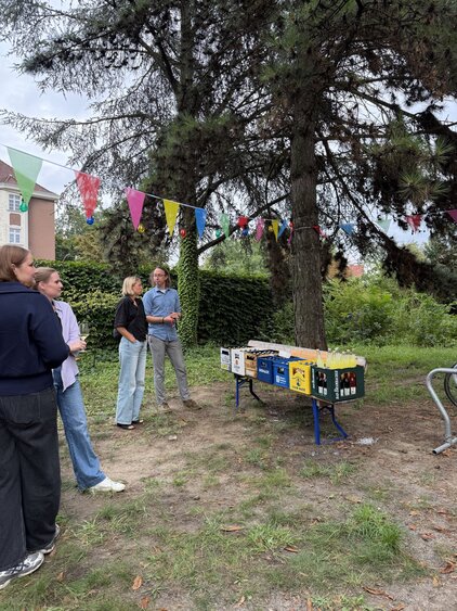 Eine Gruppe von Menschen steht zusammen unter einem Baum. Im Hintergrund befindet sich ein Tisch mit bunten Getränkekästen, und es hängen farbige Wimpel im Freien. Die Atmosphäre wirkt entspannt und gesellig.