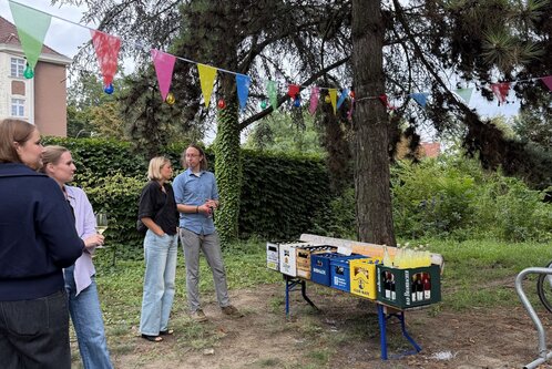 Eine Gruppe von Menschen steht zusammen unter einem Baum. Im Hintergrund befindet sich ein Tisch mit bunten Getränkekästen, und es hängen farbige Wimpel im Freien. Die Atmosphäre wirkt entspannt und gesellig.