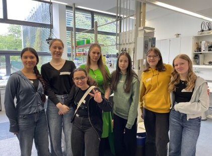 Eine Gruppe von sieben jungen Frauen steht zusammen in einem Labor. Sie lächeln freundlich in die Kamera und tragen unterschiedliche Freizeitkleidung. Im Hintergrund sind Fenster und laboreingestellte Ausstattung sichtbar.