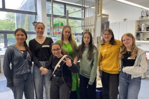 Eine Gruppe von sieben jungen Frauen steht zusammen in einem Labor. Sie lächeln freundlich in die Kamera und tragen unterschiedliche Freizeitkleidung. Im Hintergrund sind Fenster und laboreingestellte Ausstattung sichtbar.