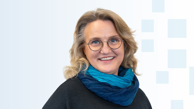 Porträt einer Frau mit blonden, lockigen Haaren und runden Brillen. Sie trägt einen dunkelblauen Schal und ein schwarzes Oberteil. Die Frau lächelt freundlich vor einem hellen, strukturierten Hintergrund.