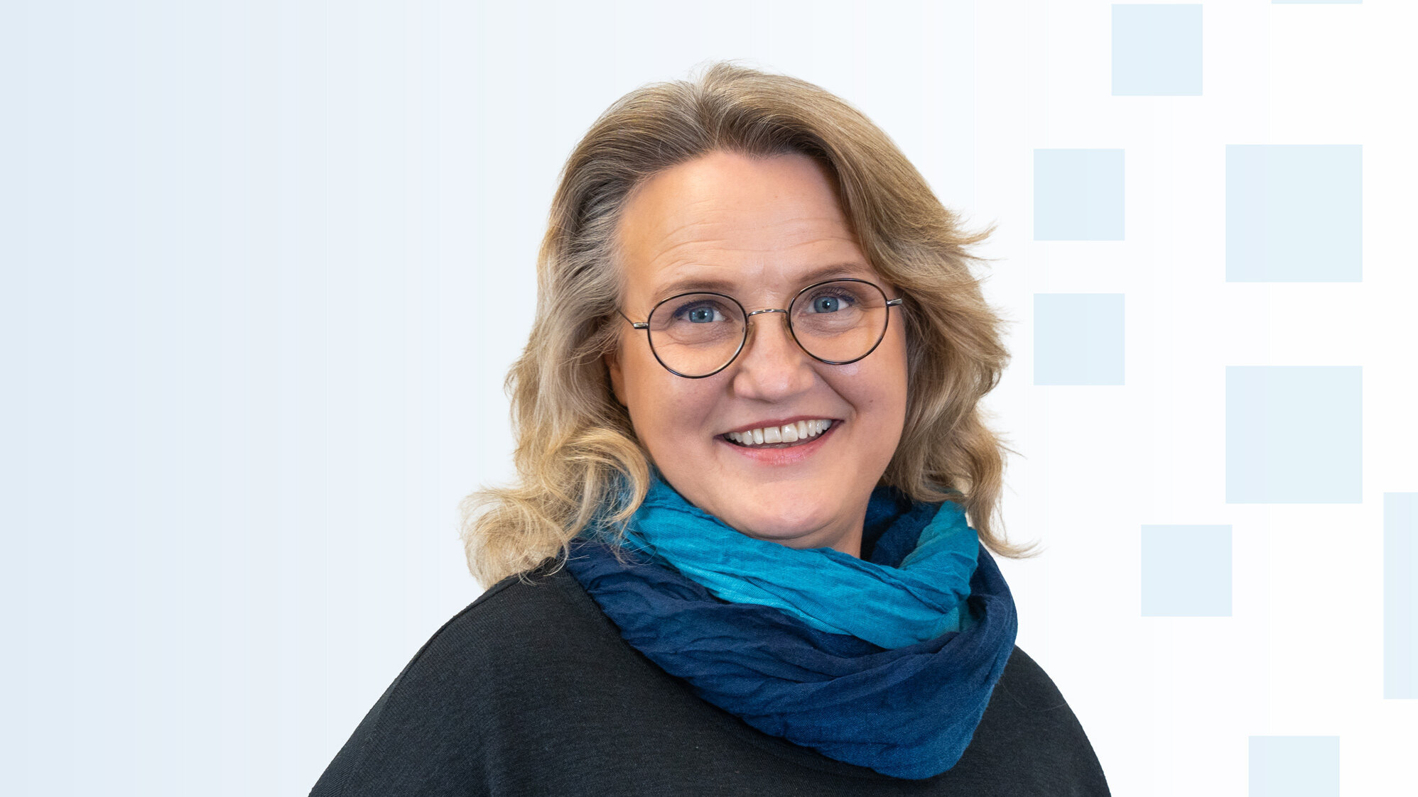 Porträt einer Frau mit blonden, lockigen Haaren und runden Brillen. Sie trägt einen dunkelblauen Schal und ein schwarzes Oberteil. Die Frau lächelt freundlich vor einem hellen, strukturierten Hintergrund.