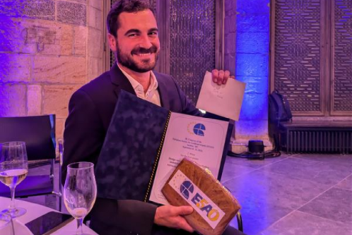 Ein Mann hält lächelnd ein Urkunde in der einen Hand und eine kleine Box in der anderen. Am Tisch neben ihm stehen ein Glas Sekt und eine Flasche. Der Hintergrund ist mit sanftem Licht beleuchtet, was eine feierliche Atmosphäre schafft.