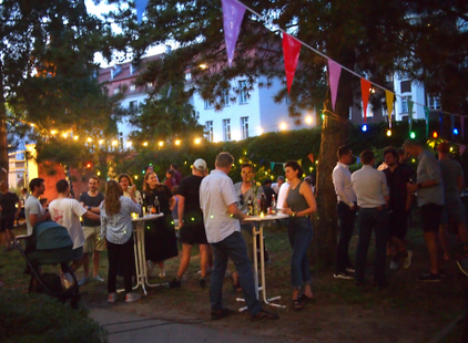 Eine fröhliche Feier im Freien, bei Nacht. Menschen stehen an Stehtischen, unter Lichtern und bunten Fähnchen. Einige unterhalten sich, andere halten Getränke. Der Hintergrund zeigt eine grüne Landschaft mit Gebäuden, die eine entspannte Atmosphäre schaffen.