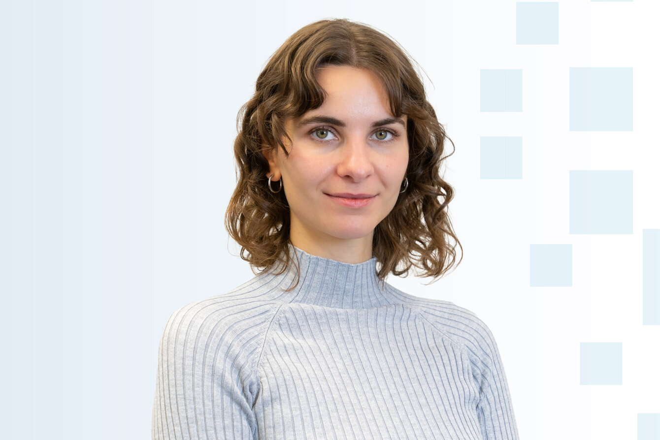 Porträt einer lächelnden Frau mit lockigem, braunem Haar, die ein hellgraues, geripptes Oberteil trägt. Der Hintergrund ist in sanften, hellen Farben mit geometrischen Mustern gestaltet. Die Frau schaut direkt in die Kamera und vermittelt eine freundliche Ausstrahlung.